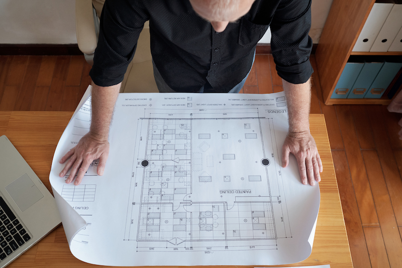 High angle view of unrecognizable mature man looking at large blueprint of ceiling while standing near office desk