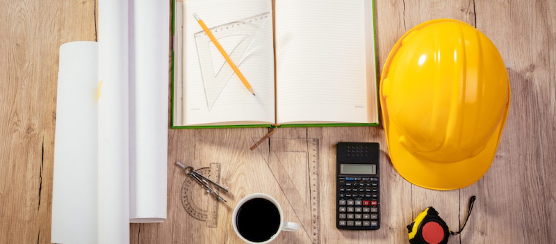 Top view of a architectonic workspace with laptop and yellow helmet. Different accessories, plans and cup of coffee is on the table.